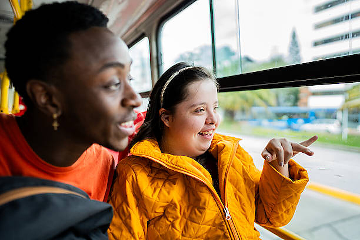 A young woman with Down syndrome smiling and pointing out the window on a bus ride with a friend